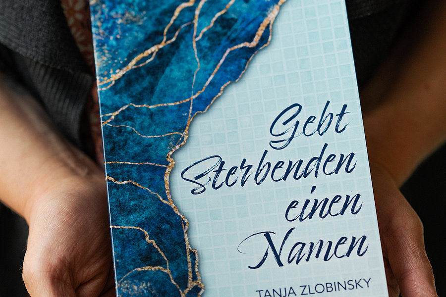 Hände halten ein Buch mit blau-weißem Marmormuster und dem Titel 'Gebt Sterbenden einen Namen' von Tanja Zlobinsky