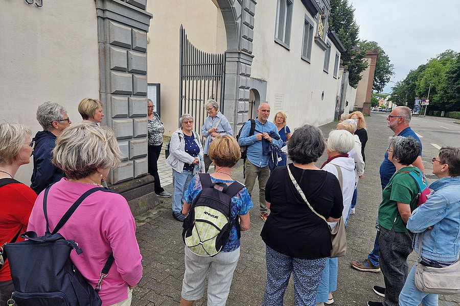 Gruppe von Menschen steht vor einem historischen Gebäude mit Torbogen und Gittertor auf einem Gehweg.