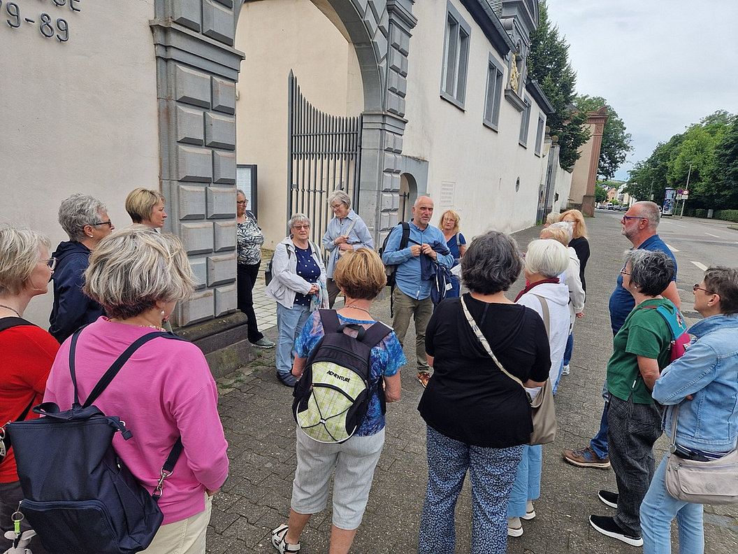 Gruppe von Menschen steht vor einem historischen Gebäude mit Torbogen und Gittertor auf einem Gehweg.