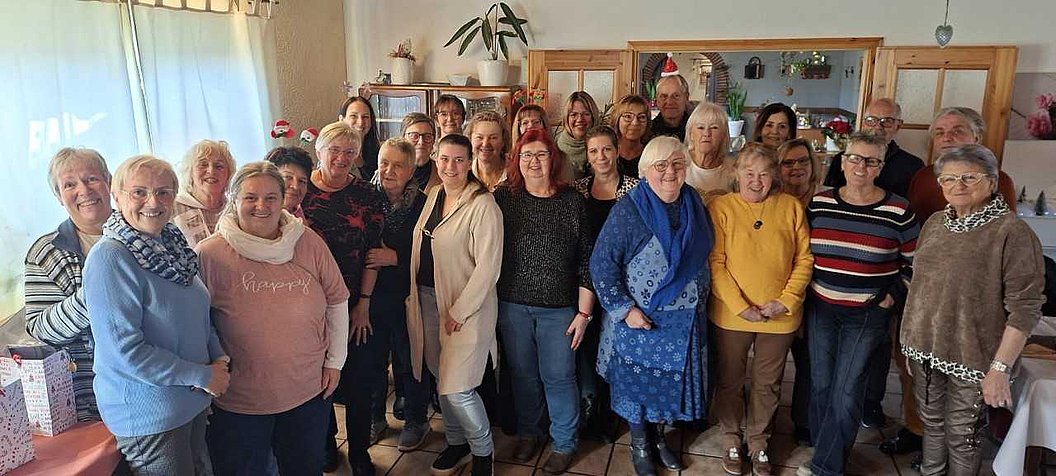 Gruppe von Menschen unterschiedlichen Alters in einem Raum mit Holzmöbeln und Pflanzen im Hintergrund.