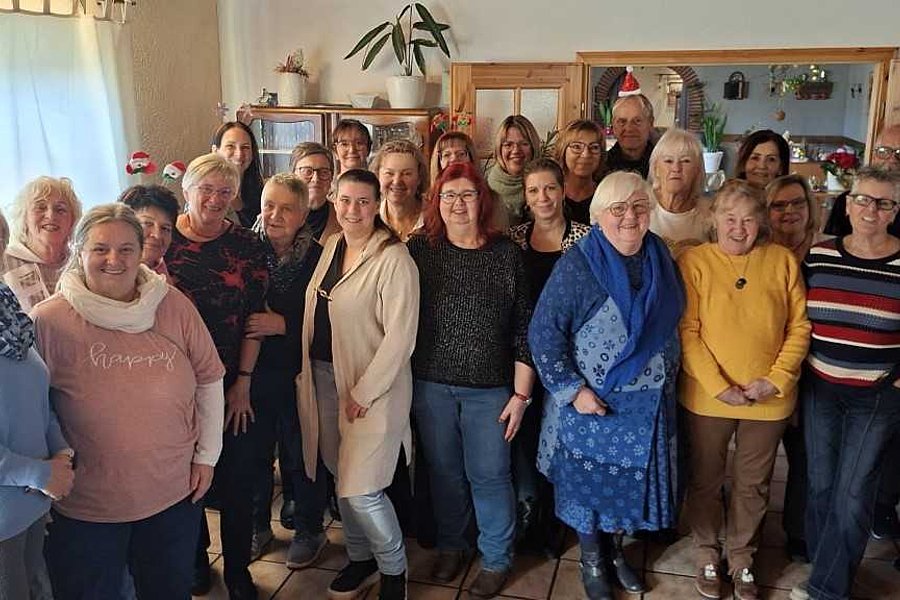 Gruppe von Menschen unterschiedlichen Alters in einem Raum mit Holzmöbeln und Pflanzen im Hintergrund.
