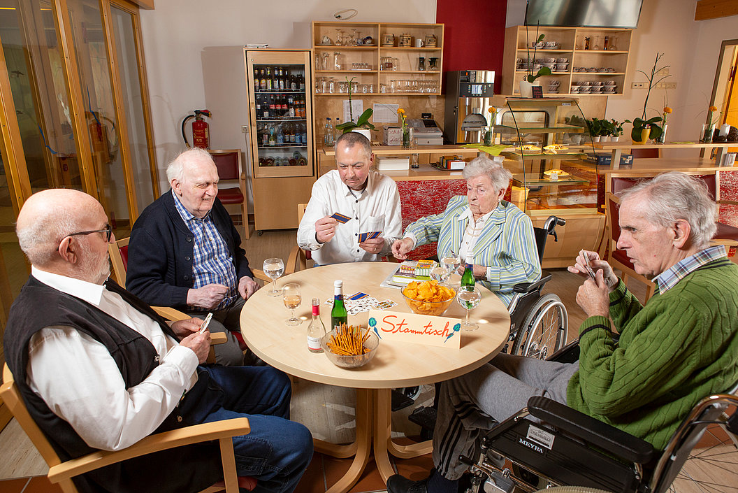 Fünf ältere Menschen sitzen um einen runden Tisch in einem Gemeinschaftsraum, spielen Karten und haben Snacks und Getränke auf dem Tisch