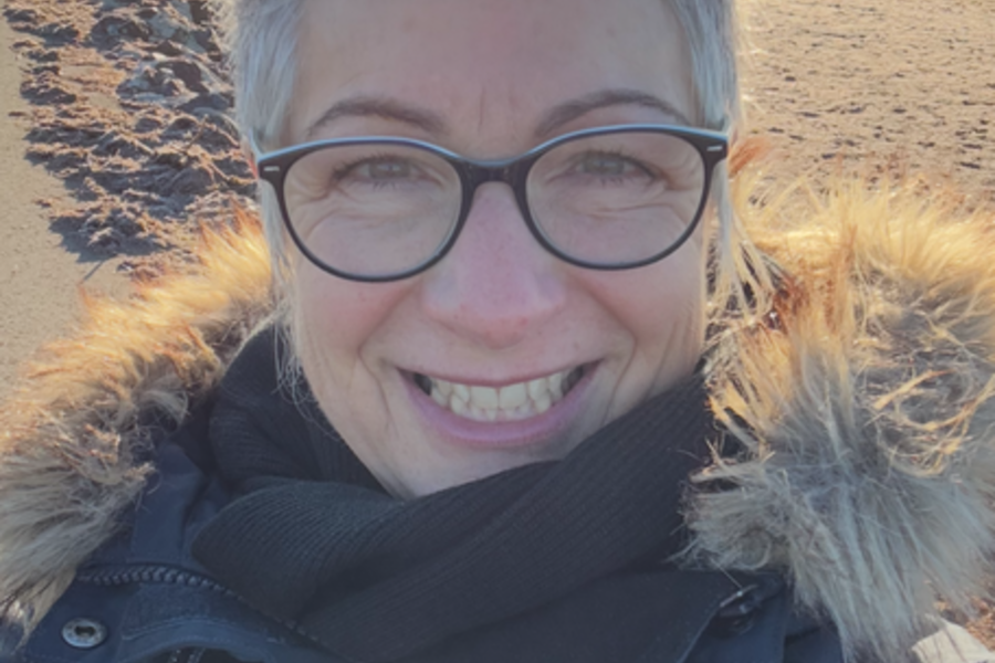 Angela Axmann mit dunklem Wintermantel mit Fellkapuze vor einer weiten, sonnigen Sandlandschaft.
