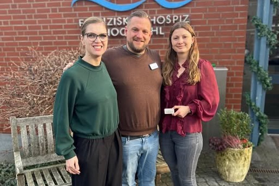 Gruppenfoto vor einem Hospiz mit drei Personen, die freundlich in die Kamera lächeln.