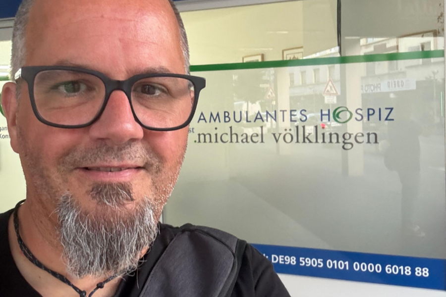 Oberkörper einer Person mit grauen Haaren und schwarzem T-Shirt vor einer Glaswand mit der Aufschrift 'AMBULANTES HOSPIZ'