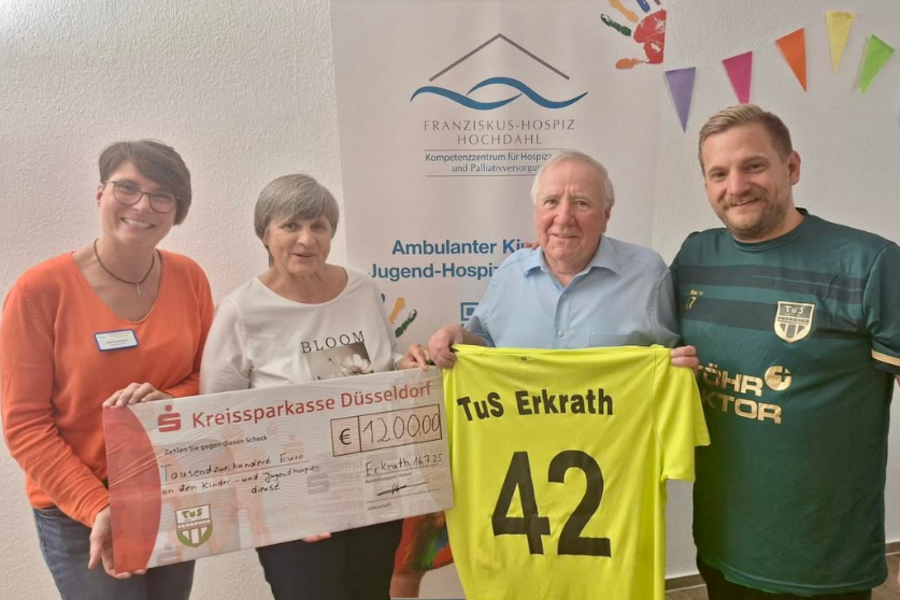 Vier Personen stehen vor einem Banner des Franziskus-Hospiz Hochdahl. Zwei halten einen großen Scheck über 12.000 Euro, zwei weitere halten ein gelbes Fußballtrikot mit der Aufschrift 'TuS Erkrath 42'.
