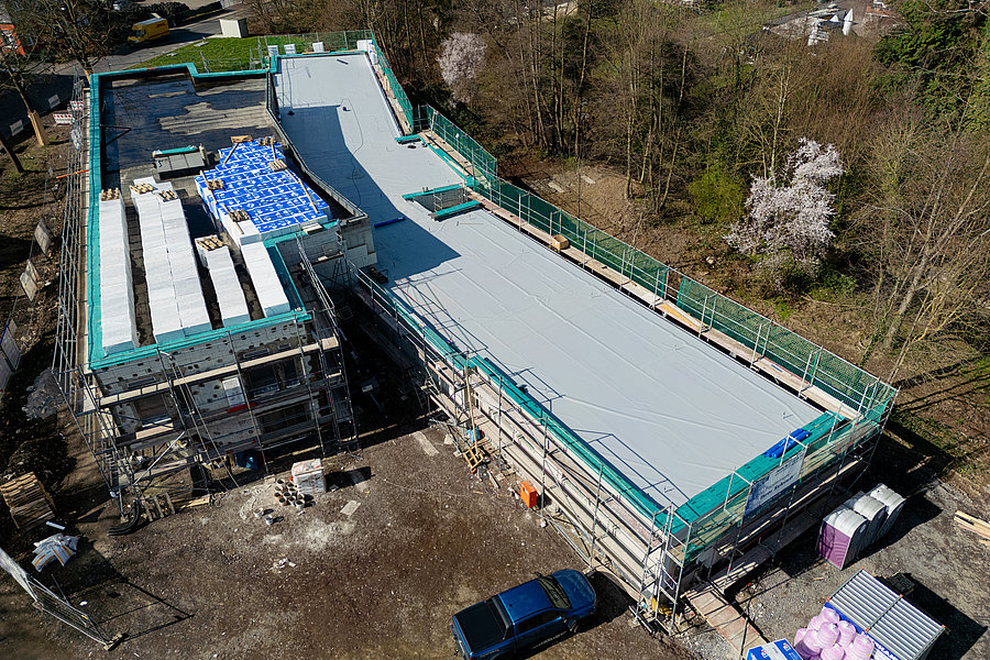 Luftaufnahme einer Baustelle mit einem langgestreckten Gebäude, das teilweise mit Gerüst und blauer Folie bedeckt ist.