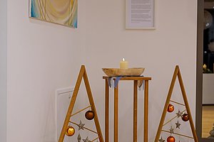 Dreieckige Holzrahmen mit Weihnachtskugeln und Sternen, dazwischen ein hoher Holzständer mit einer brennenden Kerze auf einem runden Teppich auf Holzboden vor weißer Wand mit zwei Bildern.