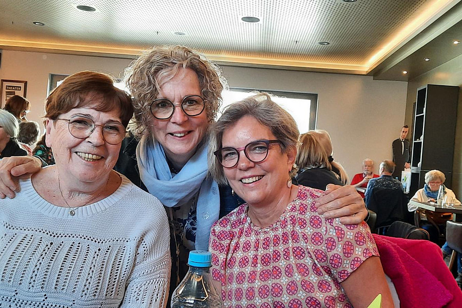 Drei Frauen sitzen an einem Tisch in einem Restaurant, zwei sitzen, eine steht dahinter und umarmt die anderen. Im Hintergrund weitere Gäste und Wanddekoration.