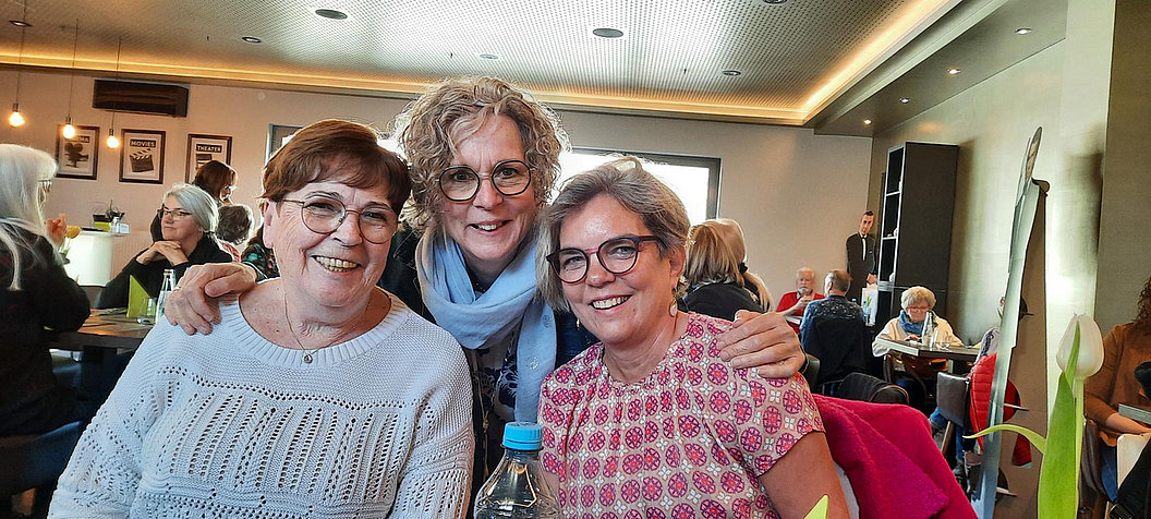 Drei Frauen sitzen an einem Tisch in einem Restaurant, zwei sitzen, eine steht dahinter und umarmt die anderen. Im Hintergrund weitere Gäste und Wanddekoration.