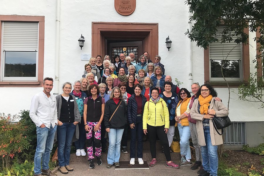 Gruppe von Menschen vor einem Gebäude mit Wappen über dem Eingang, umgeben von Fenstern und Pflanzen.