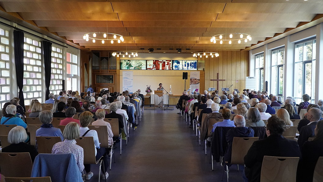 Großer Saal mit Publikum, das auf eine Bühne mit drei Personen und mehreren Bildschirmen blickt, helle Holzdecke und große Fenster an der rechten Seite.