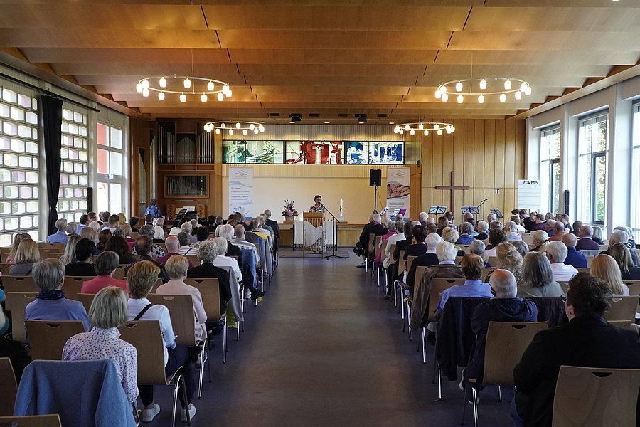 Großer Saal mit Publikum, das auf eine Bühne mit drei Personen und mehreren Bildschirmen blickt, helle Holzdecke und große Fenster an der rechten Seite.