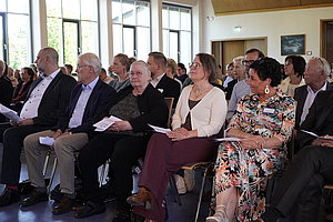 Mehrere Personen sitzen in einer Reihe auf Stühlen in einem hellen Raum mit großen Fenstern und halten Papiere in den Händen.