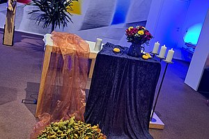 Kreuz mit Jesusfigur, daneben ein mit Stoff bedeckter Tisch mit Kerzen und Blumen, davor zwei runde Arrangements mit Pflanzen und kleinen Kerzen auf dem Boden.