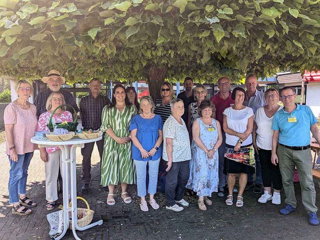 Gruppe von 15 Personen steht unter einem großen Baum, einige tragen sommerliche Kleidung, im Vordergrund ein weißer Stehtisch mit zwei Körben.