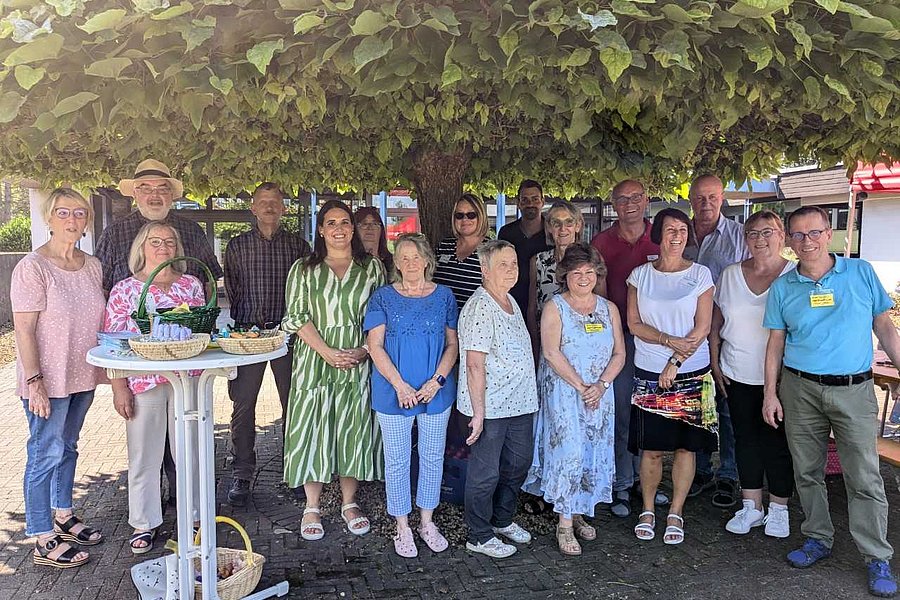 Gruppe von 15 Personen steht unter einem großen Baum, einige tragen sommerliche Kleidung, im Vordergrund ein weißer Stehtisch mit zwei Körben.