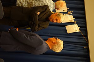 Drei ältere Personen stehen vor einer blauen Vorhangwand und unterhalten sich, im Hintergrund hängen mit Klammern befestigte Papiere an einer Leine