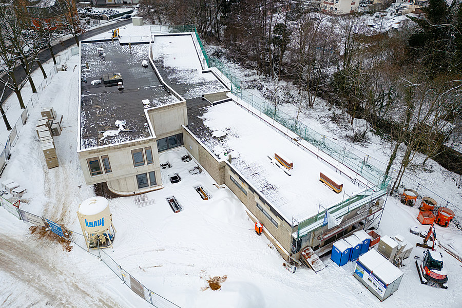 Luftaufnahme einer schneebedeckten Baustelle mit zwei Gebäuden, Baugeräten, Containern und einem Zaun.