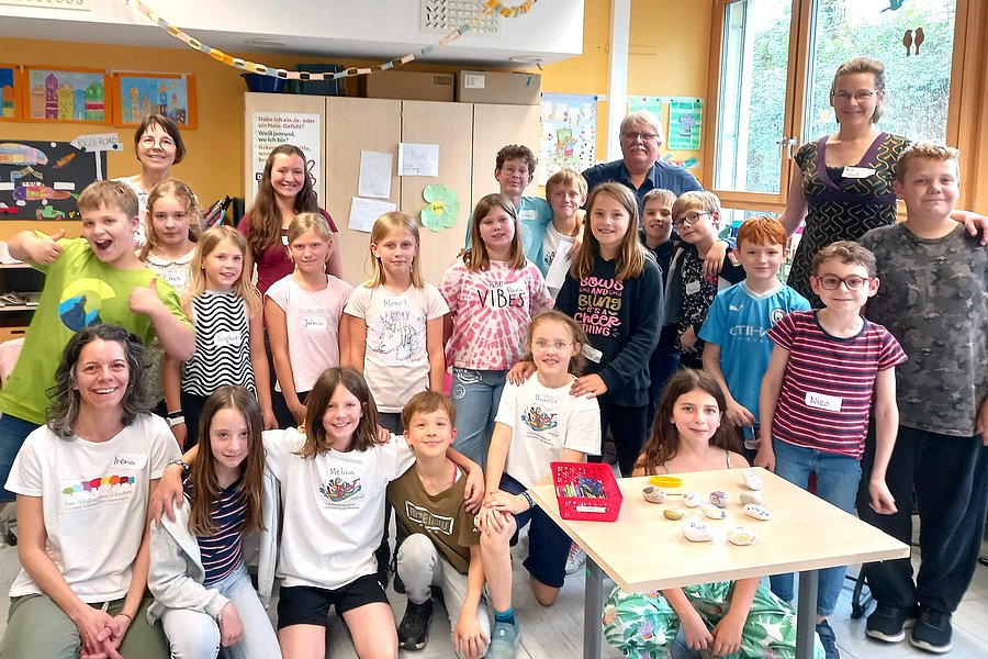 Gruppe von Kindern und einer erwachsenen Person in einem Klassenzimmer, einige sitzen, andere stehen, Tisch mit kleinen Gegenständen vorne rechts.
