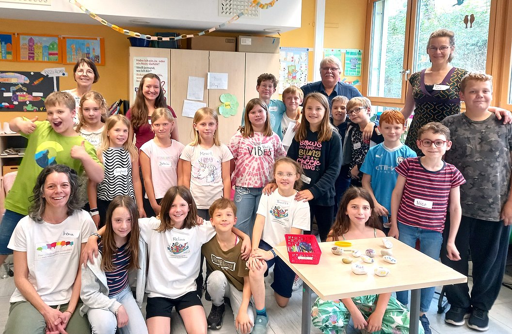 Gruppe von Kindern und einer erwachsenen Person in einem Klassenzimmer, einige sitzen, andere stehen, Tisch mit kleinen Gegenständen vorne rechts.