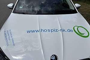 Vorderansicht einer weißen Skoda-Autohauptmotorhaube mit der blauen Webadresse 'www.hospiz-nk.de' und einem grünen Logo rechts daneben.