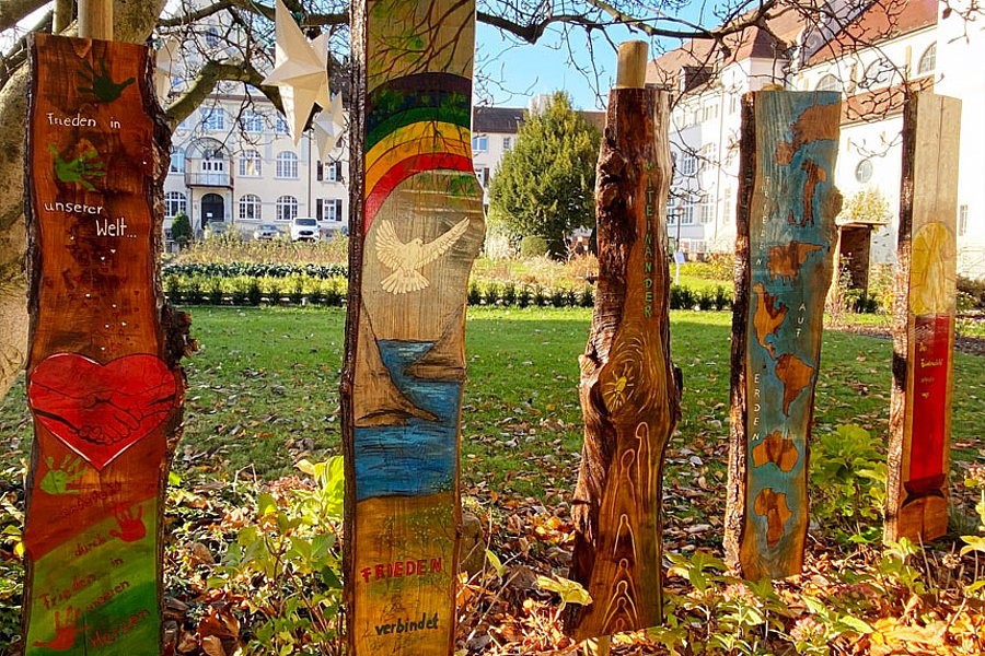 Fünf bemalte Holzpfähle mit Friedensthemen, darunter Herz, Taube, Regenbogen und Schrift, stehen im Herbstlaub auf einer Wiese vor Gebäuden.