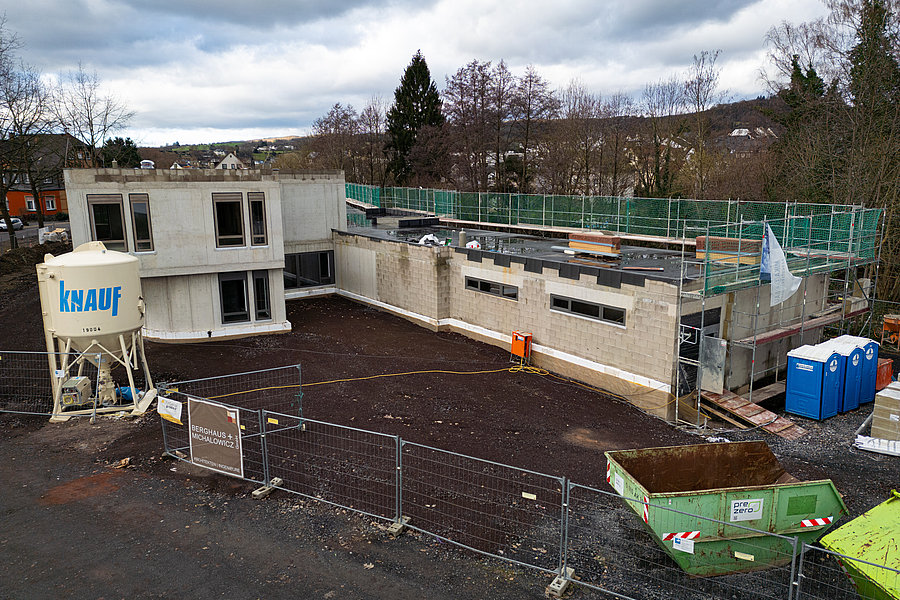 Baustelle mit teilweise errichtetem zweistöckigem Gebäude, Baustoffbehälter mit Aufschrift 'knauf', Bauzaun und zwei blauen Toilettencontainern.