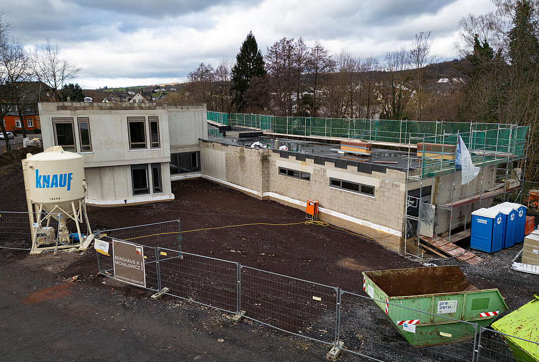 Baustelle mit teilweise errichtetem zweistöckigem Gebäude, Baustoffbehälter mit Aufschrift 'knauf', Bauzaun und zwei blauen Toilettencontainern.