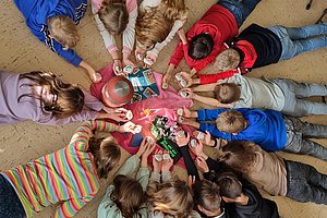 Kinder liegen im Kreis auf dem Boden und halten ihre Hände auf ein Spielbrett in der Mitte