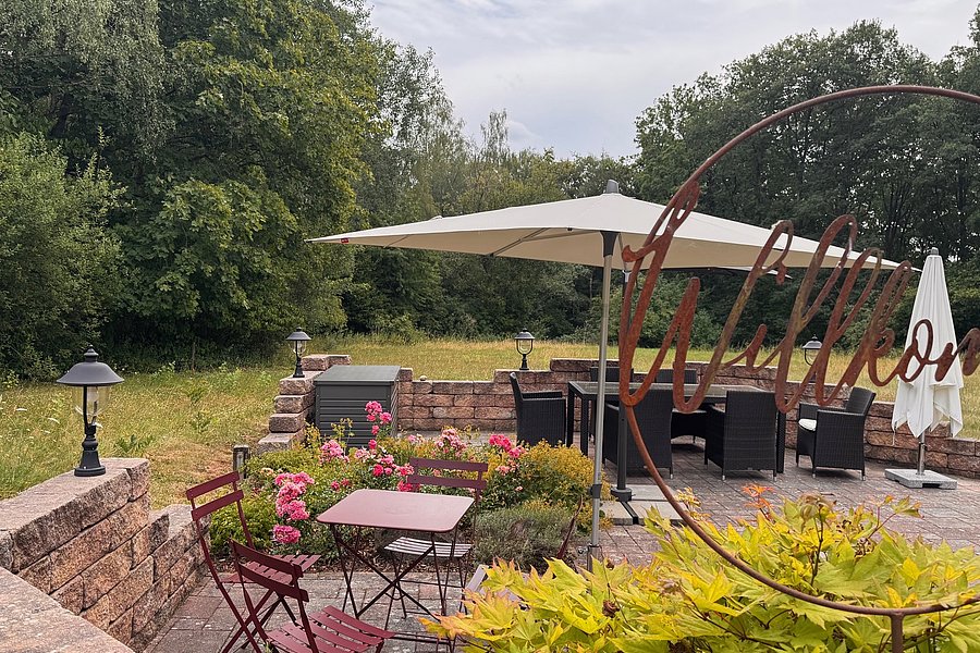Terrasse mit Tischen, Stühlen, Sonnenschirmen und Pflanzen vor einem Wald