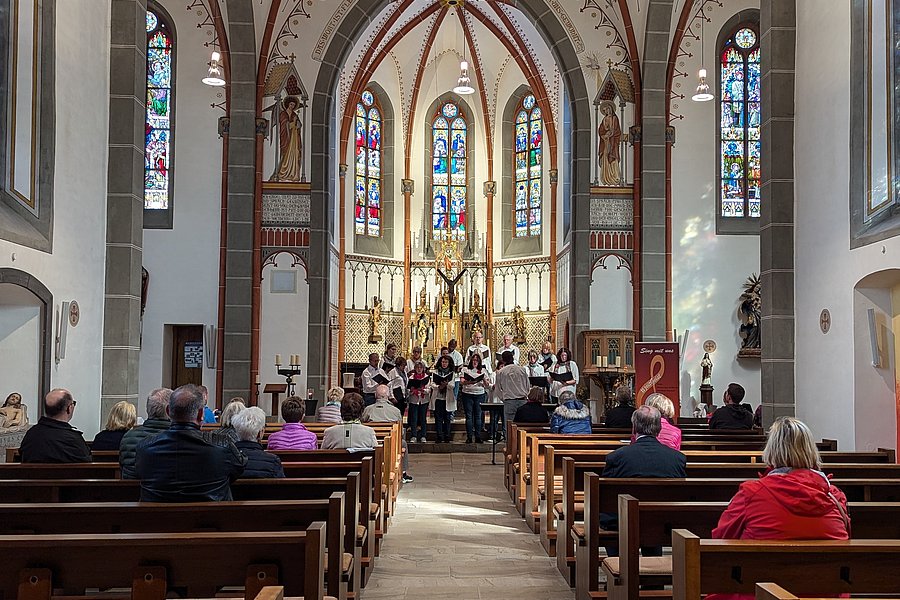 Innenansicht einer Kirche mit bunten Glasfenstern, Altar und einer Gruppe singender Menschen vor der Gemeinde.