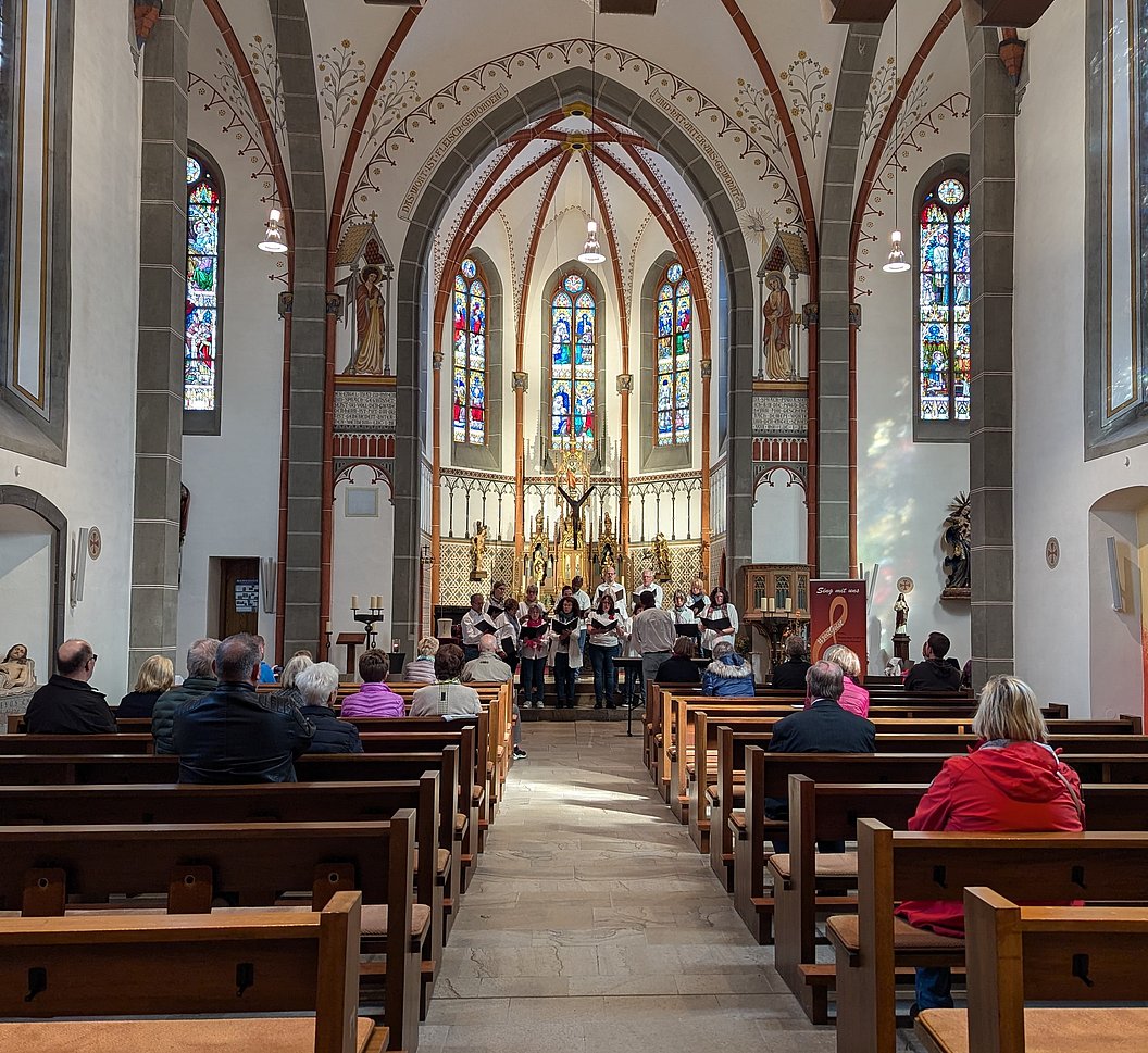 Innenansicht einer Kirche mit bunten Glasfenstern, Altar und einer Gruppe singender Menschen vor der Gemeinde.