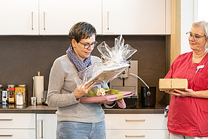 Zwei Frauen in einer Küche, eine hält ein Geschenkpaket in Folie und Küchenhandschuhe, die andere ein kleines Geschenkpaket.