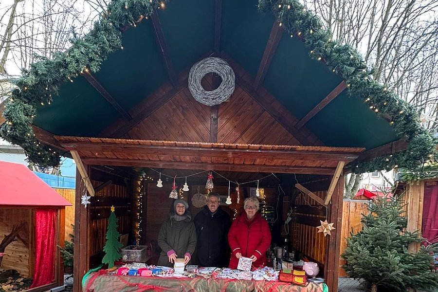 Weihnachtlich geschmückte Holzhütte mit drei Personen hinter einem mit Tüchern bedeckten Verkaufstisch auf gepflastertem Platz.