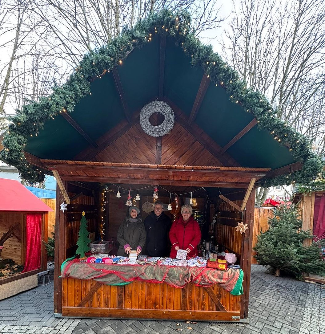 Weihnachtlich geschmückte Holzhütte mit drei Personen hinter einem mit Tüchern bedeckten Verkaufstisch auf gepflastertem Platz.