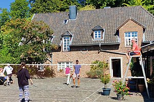 Mehrere Personen spielen Badminton auf einem gepflasterten Hof vor einem zweistöckigen Backsteinhaus mit Dachfenstern und Schornstein.