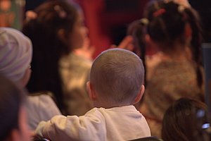 Rückenansicht eines Kindes mit Glatze in einem Rollstuhl inmitten einer Gruppe von Kindern in einem Innenraum