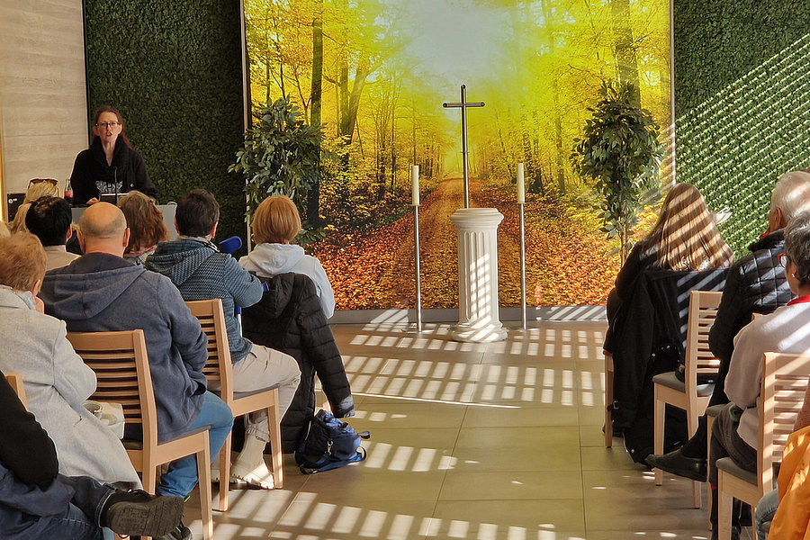 Mehrere Personen sitzen auf Holzstühlen in einem Raum mit grünem Pflanzenwand-Dekor und einem großen Wandbild eines herbstlichen Waldes mit Kreuz auf einem weißen Podest.
