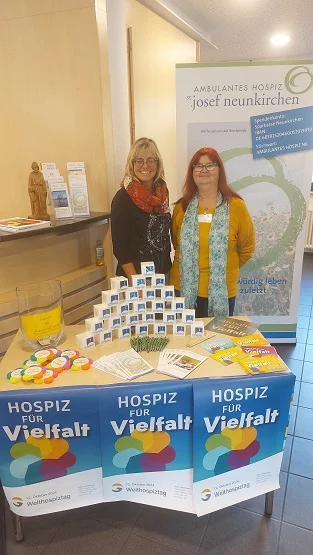 Zwei Personen stehen hinter einem Tisch mit Informationsmaterial und Werbeartikeln, darauf drei Plakate mit dem Text 'Hospiz für Vielfalt' und einem bunten Herzsymbol.