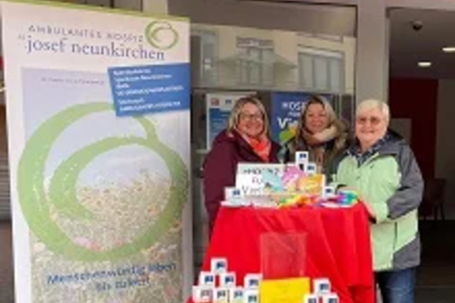 Drei Personen stehen hinter einem mit rotem Tuch bedeckten Tisch mit gestapelten weißen Würfeln und Informationsmaterial vor einem Gebäude mit einem Banner der Ambulanten Hospiz Josef Neunkirchen.