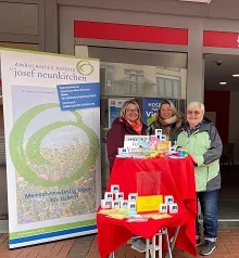 Drei Personen stehen hinter einem mit rotem Tuch bedeckten Tisch mit gestapelten weißen Würfeln und Informationsmaterial vor einem Gebäude mit einem Banner der Ambulanten Hospiz Josef Neunkirchen.