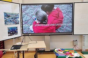 Bildschirm zeigt eine ältere Person und ein Kind in roten Jacken von hinten mit dem Text 'MODUL 2 VORSORGEN UND ENTSCHEIDEN' in einem Klassenraum mit Tischen und bunten Papierstücken