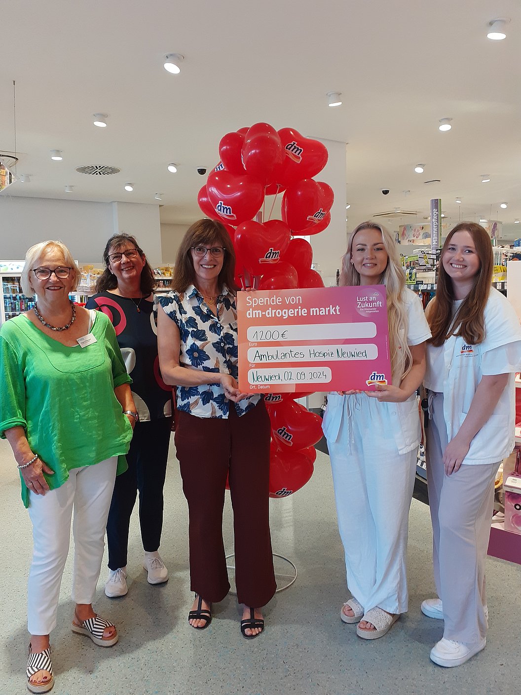 Fünf Frauen stehen in einem Geschäft, zwei halten ein großes pinkes Schild mit Text, im Hintergrund ein rotes Ballonbogen.