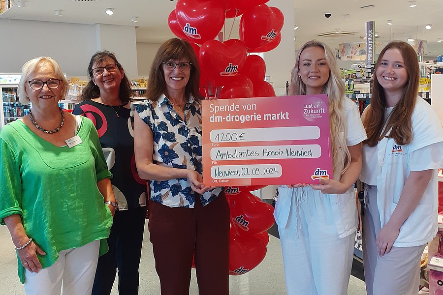 Fünf Frauen stehen in einem Geschäft, zwei halten ein großes pinkes Schild mit Text, im Hintergrund ein rotes Ballonbogen.