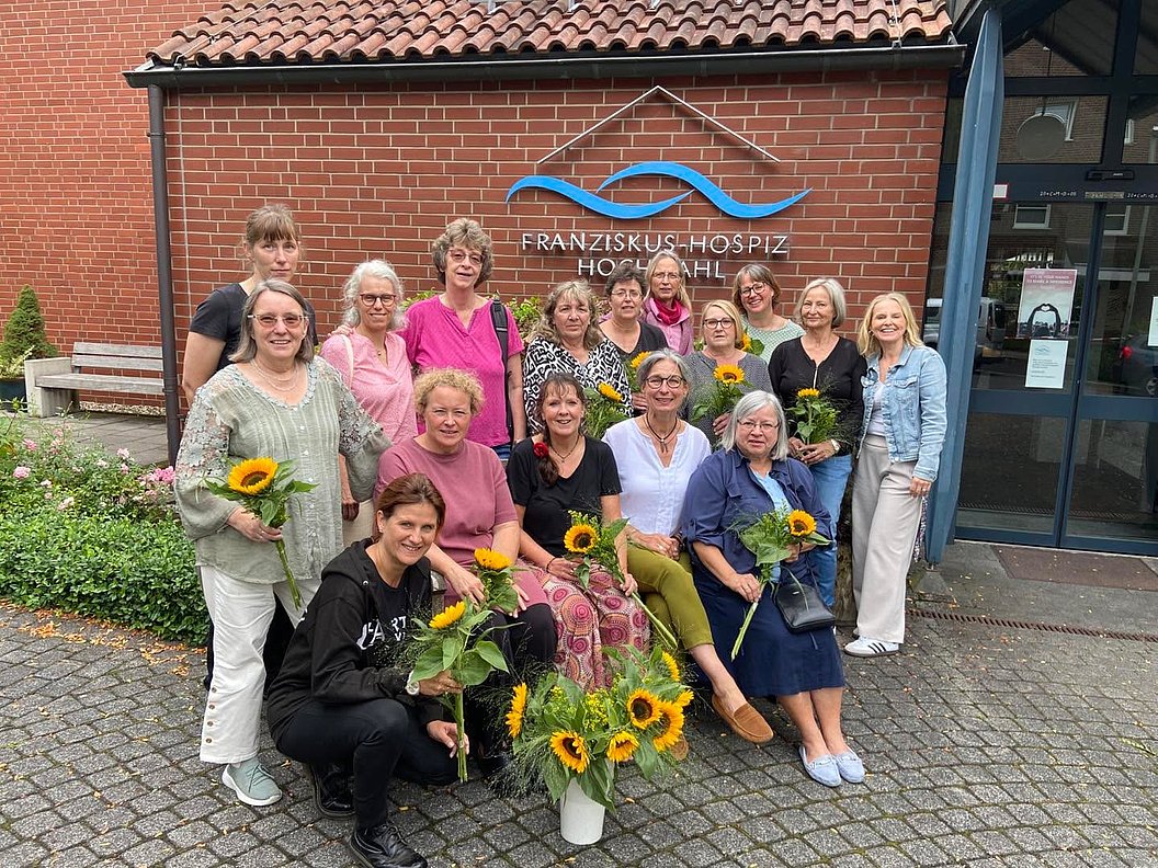 Gruppe von Menschen vor dem Gebäude des Franziskus-Hospiz Hochsauerland, einige halten Sonnenblumen, Pflanze mit Sonnenblumen steht vorn auf dem Boden.