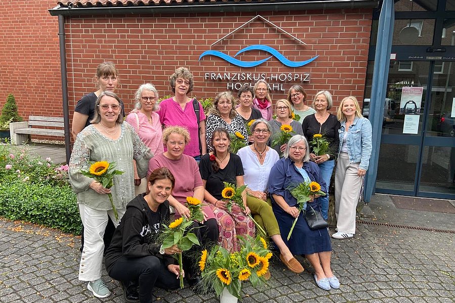 Gruppe von Menschen vor dem Gebäude des Franziskus-Hospiz Hochsauerland, einige halten Sonnenblumen, Pflanze mit Sonnenblumen steht vorn auf dem Boden.