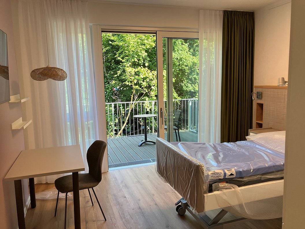 Zimmer mit Einzelbett auf Rollen, Schreibtisch mit Stuhl, Lampe und Balkon mit Blick auf grüne Bäume.
