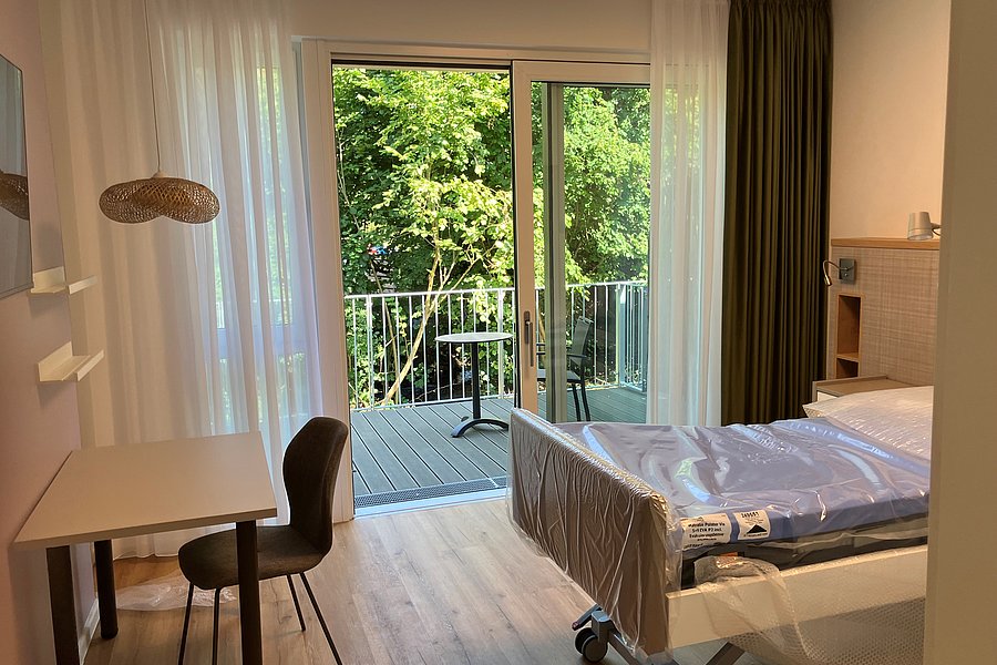 Zimmer mit Einzelbett auf Rollen, Schreibtisch mit Stuhl, Lampe und Balkon mit Blick auf grüne Bäume.