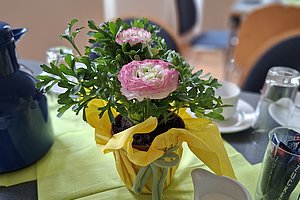 Blumengesteck mit rosa Ranunkel und grünen Blättern in gelbem Papier auf einem gedeckten Tisch mit Tassen, Gläsern und Kaffeekanne